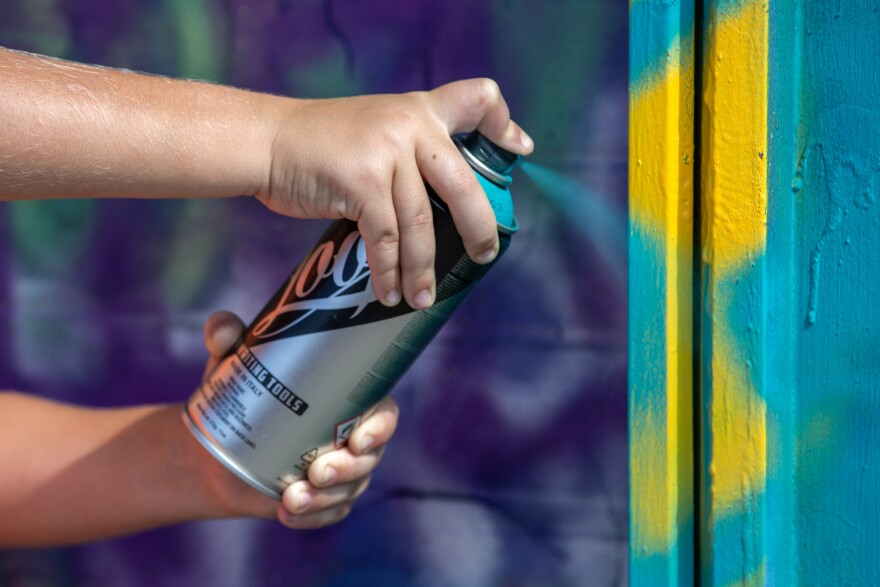 A close up of a set of hands spraying blue paint onto a blue and yellow-painted wall. 
