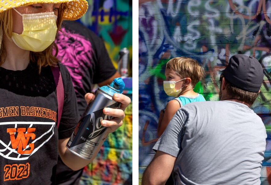 A diptych of two vertical images. The left image is of a teen girl, who is White, wearing a yellow wide brimmed hat and yellow face mask, looks at a can of blue paint with a clogged opening. The right image is of a young boy, who is White, has blonde hair and is wearing a yellow face mask, as he is held by a man in a gray t-shirt. In the background is a completely graffitied over wall. 