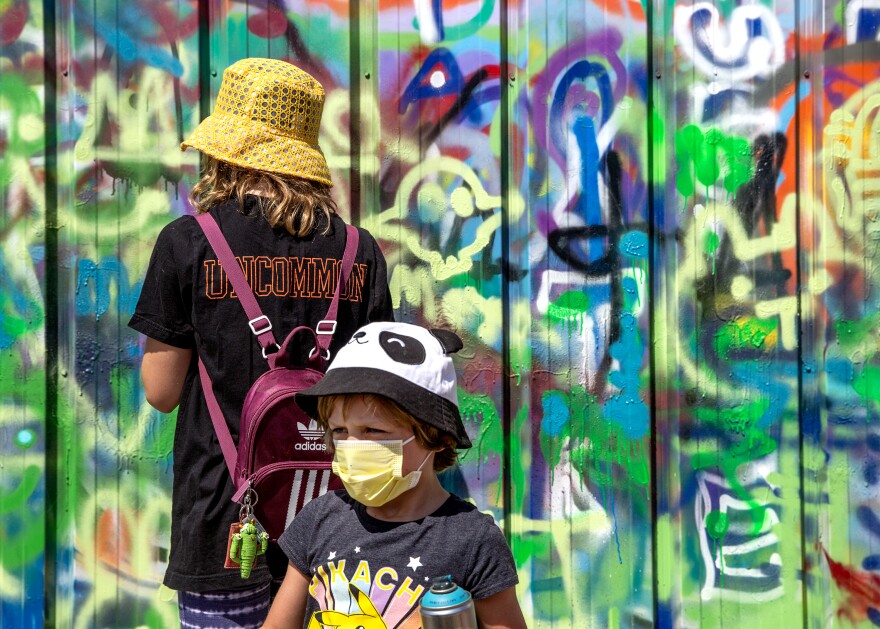 A young boy, who is White and wearing a yellow face mask and panda hat, walks past a teen girl with her back to the camera. Behind them both is a completely graffitied over wall. 