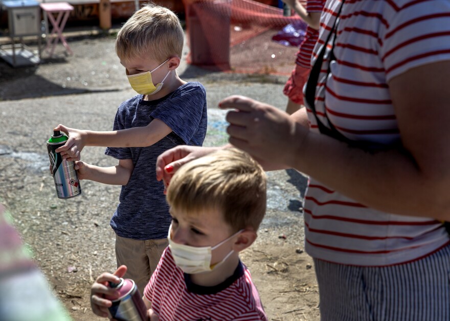 Two young boys, who are both blonde and wearing face masks, spray paint at a wall while standing next to a woman, whose head is cropped out of the frame. 