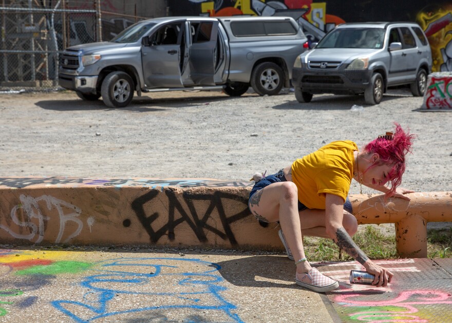 A woman, who is White, with dyed pink hair and who is wearing a bright yellow t-shirt, kneels down by a stretch of concrete and with a can of pink spray paint. Behind her, you can see a pair of gray SUVs. 