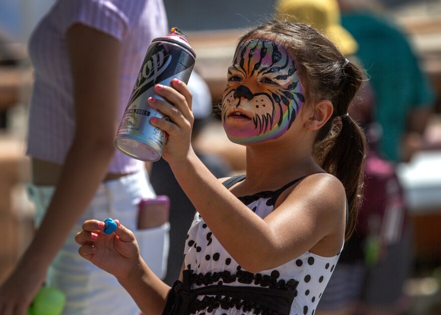 A young girl examines a can of spray paint. Her face is painted to look like a rainbow-colored tiger. 