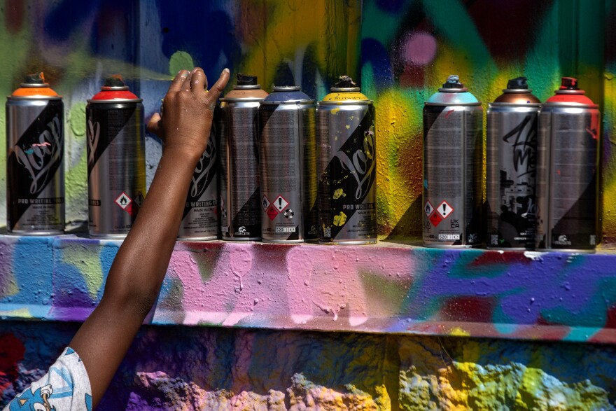 A group of spray paint cans sits in a row on a ledge, which is completely covered in spray paint. An arm, which belongs to a young Black boy, is reaching up to grab one of them. 