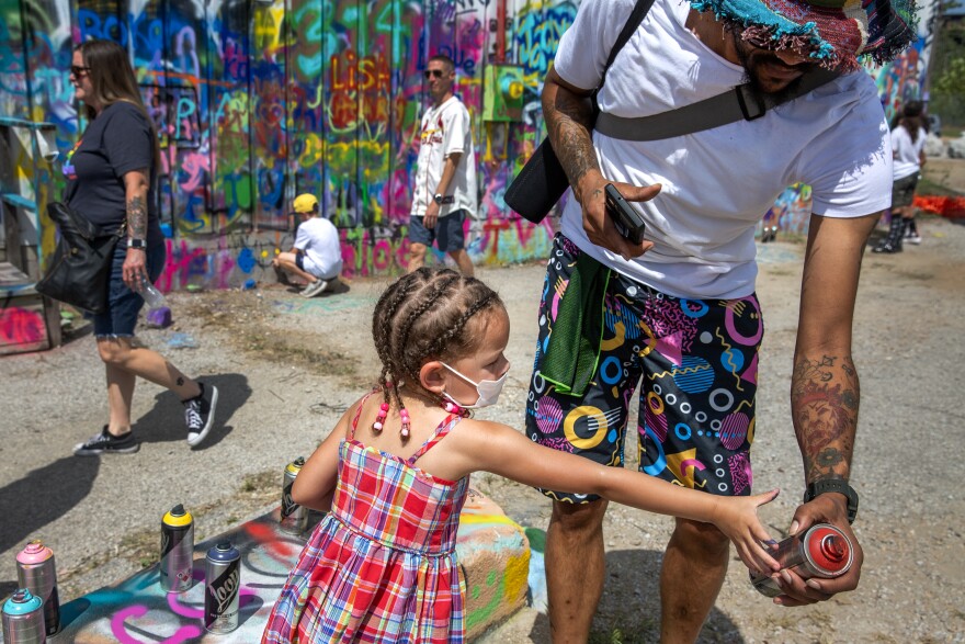 A young girl wearing a plaid red dress and whose hair is in braids, reaches of a can of spray paint being held by a man in a white t-shirt, shorts with multicolored circles on them and a bucket hat. Behind them, people walk in front of a graffiti-covered wall. 