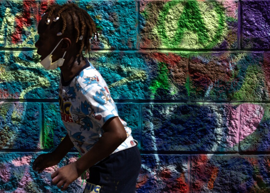 A young Black boy, wearing a yellow mask around his chin and a Sonic the Hedgehog t-shirt, runs past a graffiti-covered brick wall. 