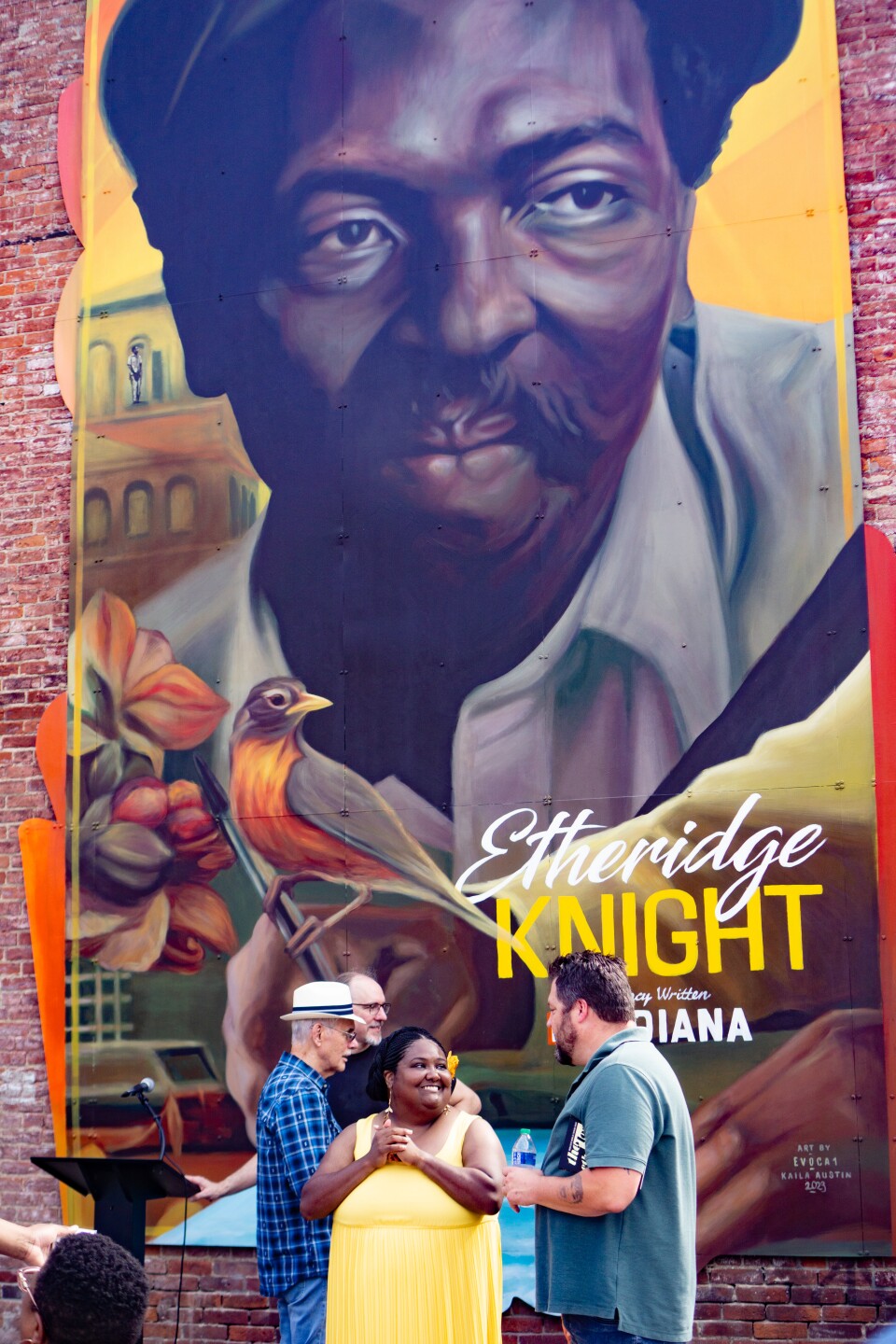 African American woman in a yellow dress talking to white man in front of a large mural of Etheridge Knight, including Knight's face and a picture of a bird.