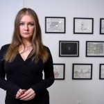 A woman wearing a black dress looks at the camera in front of eight framed drawings.
