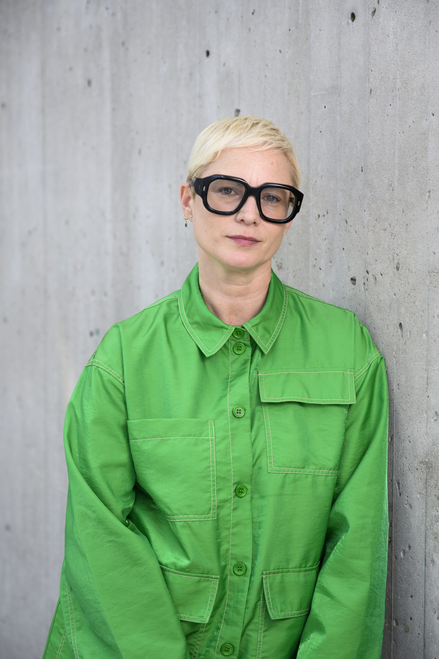 A woman in a baggy men's shirt and glasses stands in front of a wall