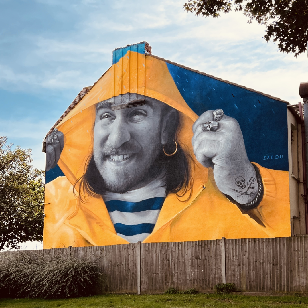 A large, very realistic painting of a man in a yellow hoodie painted on the side of a building. The man’s skin is painted grey to make the other bright colours stand out even more!
