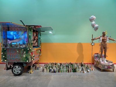 An installation showing a food cart that is dispalying a video work and has been decorated with figurines (at left), dozens of glass bottles (center), and a mannequin hooked up to medical devices standing on an arm chair (right). 
