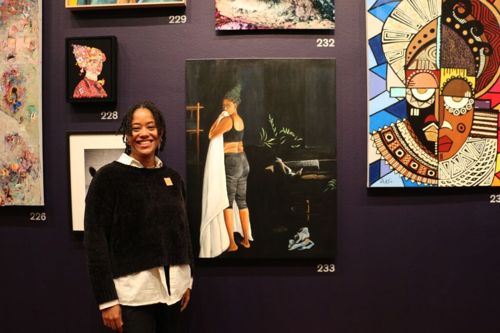 Chase Irvan stands in front of her painting. She is wearing a black sweater over a collared white shirt and black braids. The painting is of a Black woman in black yoga pants and a black sports bra, holding a white towel, with her back toward the camera and her face turned toward it, covering her mouth with the towel.