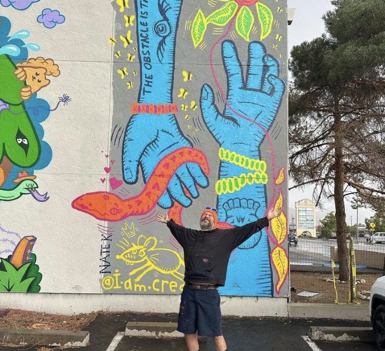 A photo of artist Nathanael Kuhn standing in front of a mural he worked on. His arms are spread wide.