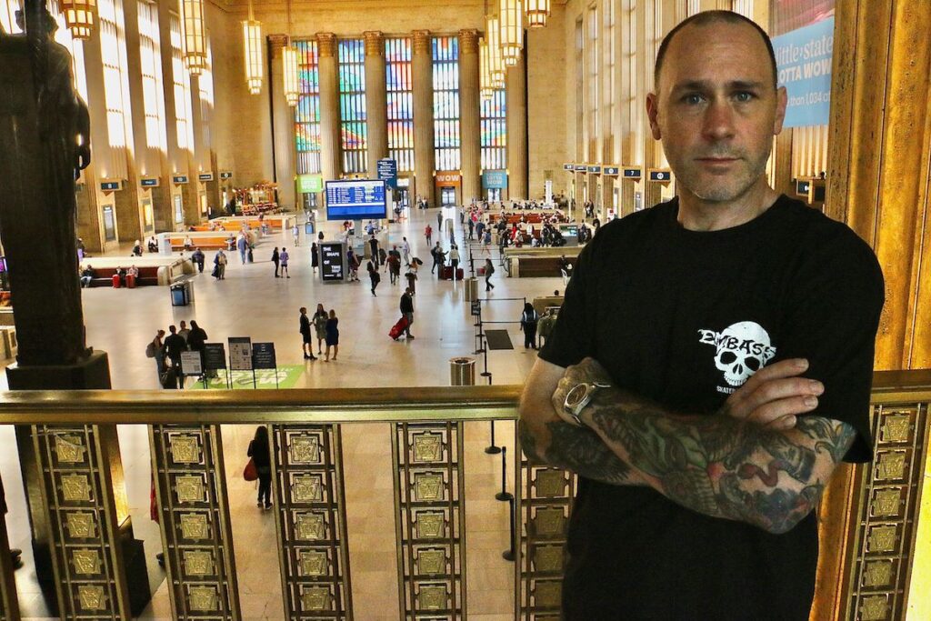 Adam Crawford poses at a railing. Behind him is the 30th Street Station main area with his artwork of rainbow-coloured vinyl on the huge windows at the front.