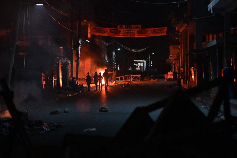 A barricaded street at night with a fire in the background an silhouettes of four people in front of it.