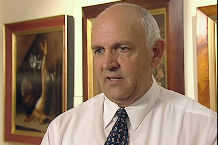 A man in a white shirt speaks in front of paintings.