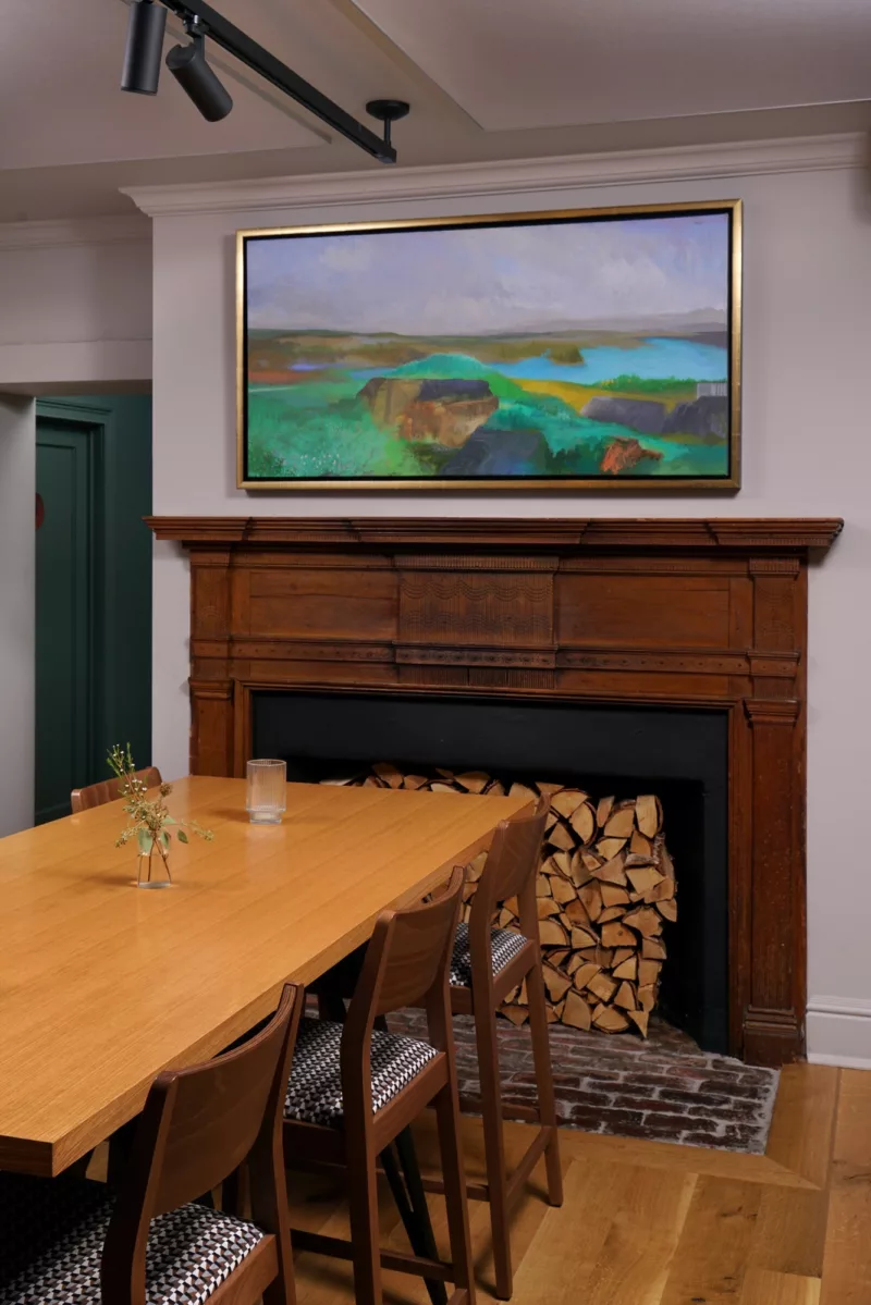 An image shows a warm and welcoming old fashioned dining room with a wood table and chairs in front of a wood framed fireplace. Above the fireplace is a colorful landscape painting by James Brantley, framed in a gold frame. 