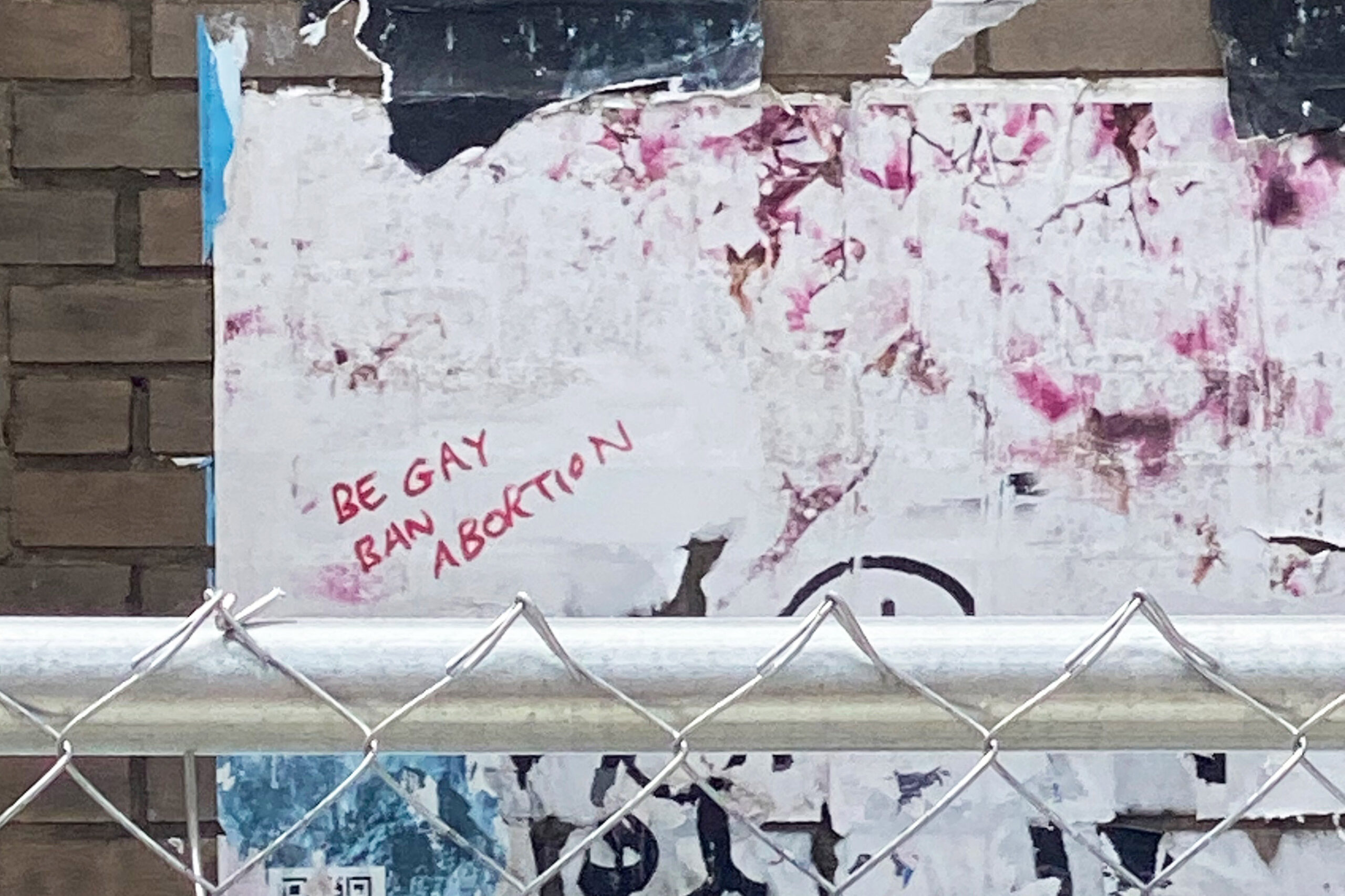 A photograph of graffiti written in red marker behind a chain-link fence. It reads, 