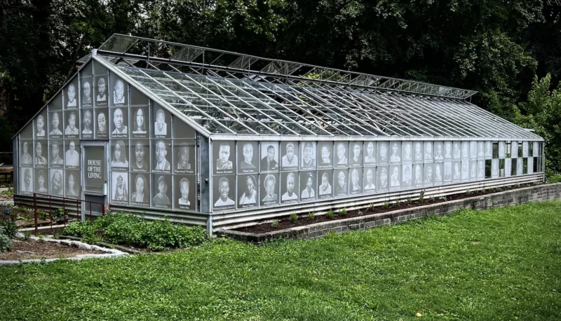 A greenhouse with a door labeled “House of the Living” is surrounded by lush grass and shrubs. On the panes of glass are large black and white photographic images of men and women and children lost to homicide in Philadelphia.