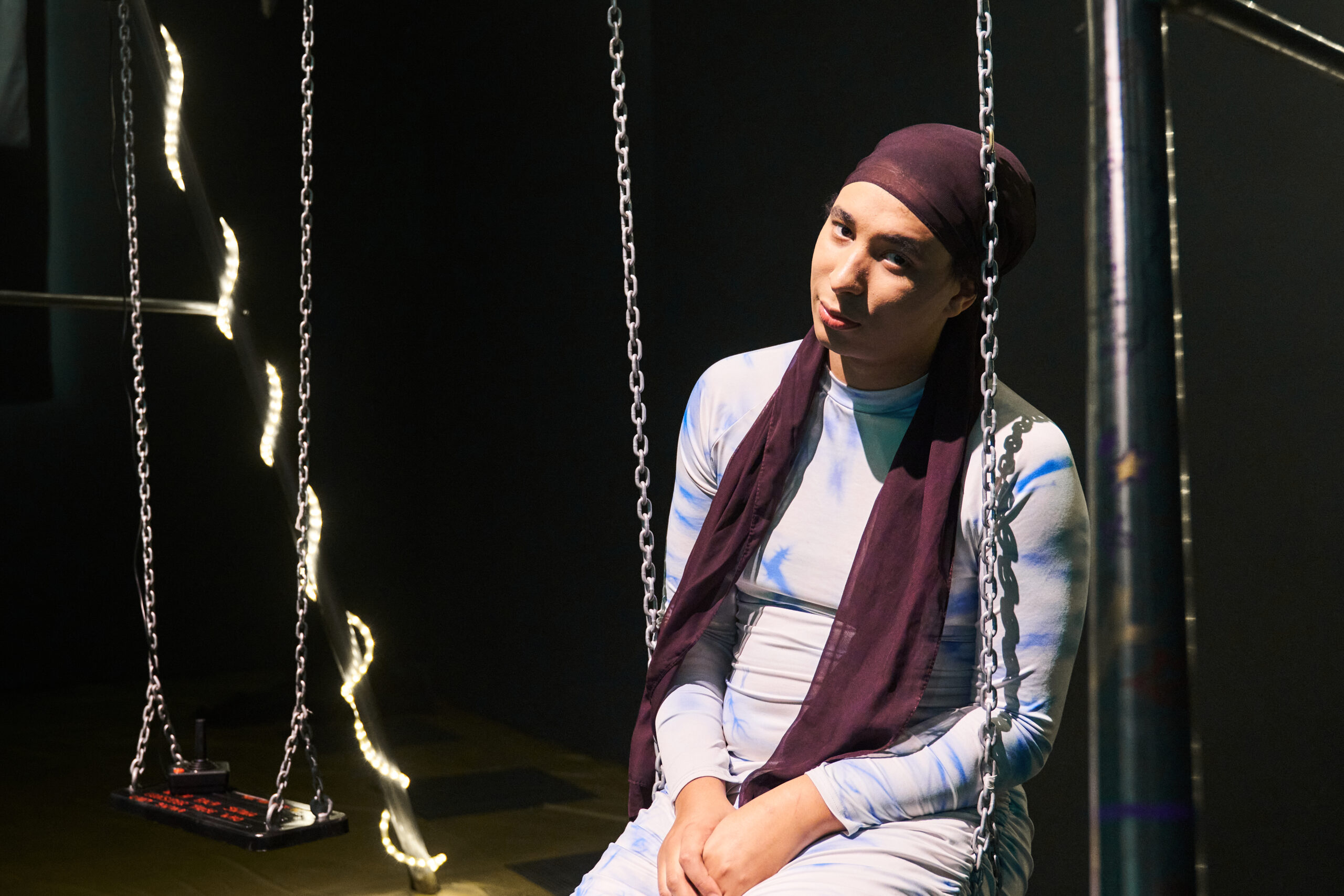 A woman wearing a headscarf sits on a swing in a dark space