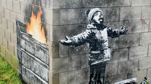 An artwork by Banksy on the side of a garage depicts a child dressed for snow playing in the falling ash and smoke from a skip fire, in Port Talbot, Wales.
