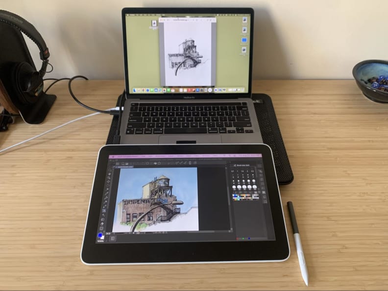 The Wacom One 13 Touch drawing tablet appears in front of a Macbook Pro on a desk with a few other desk items around it, the Wacom’s pen appears next to it.