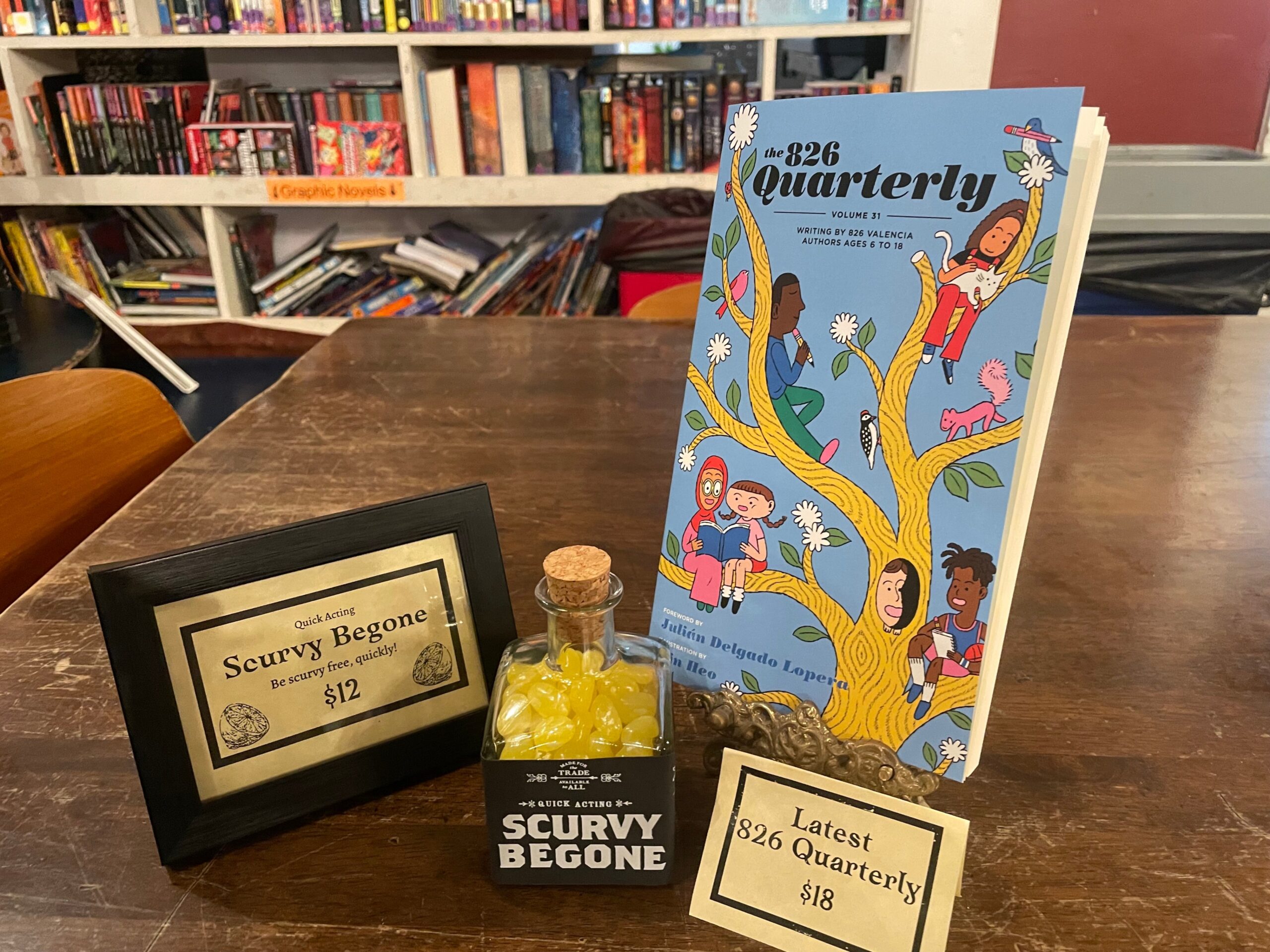 A "Scurvy Begone" bottle of candy and a children's literary journal sit on a a wooden table in a pirate-themed storefront.