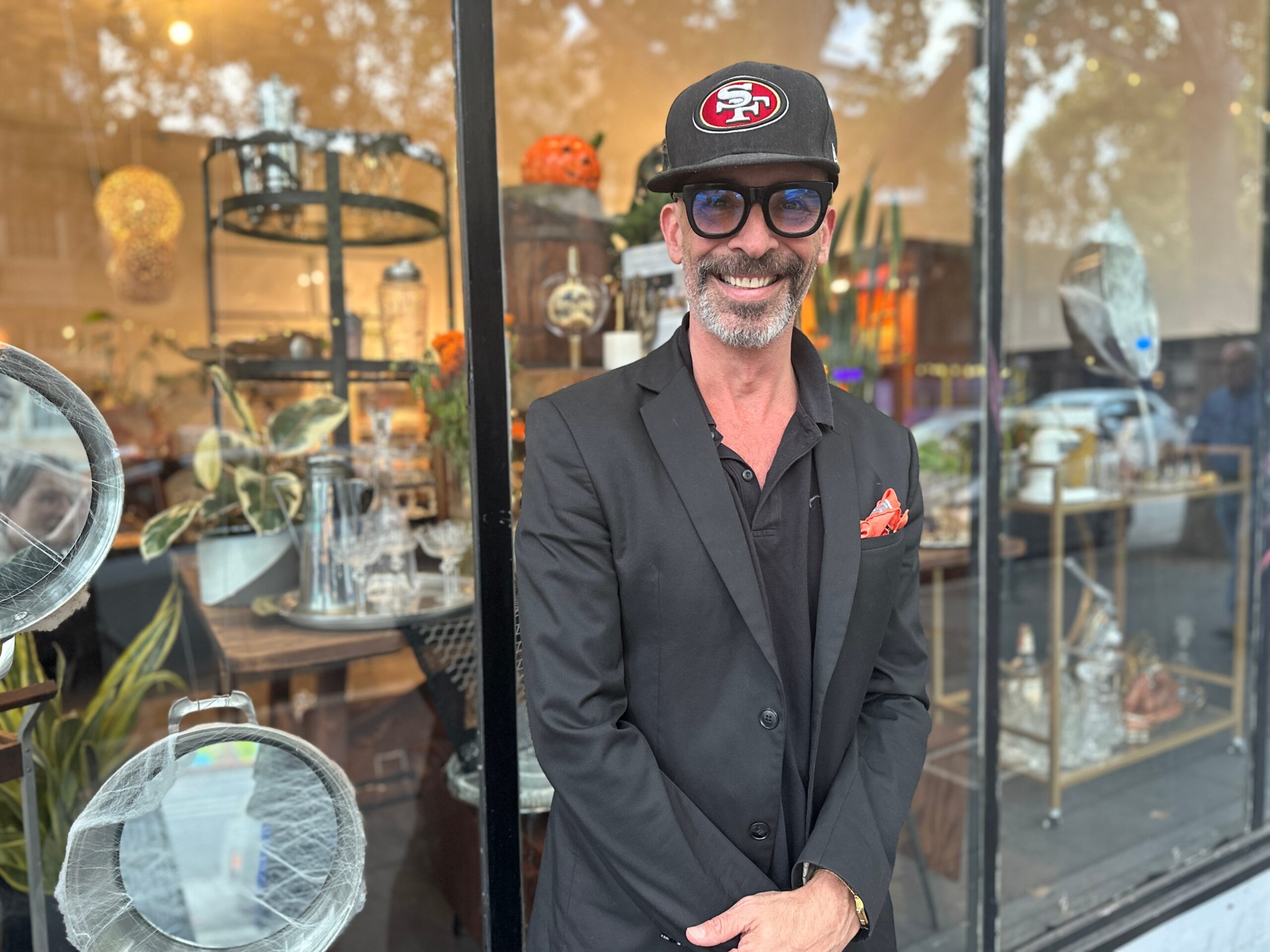 A man with an SF baseball cap stands outside a storefront filled with vintage cookware, glassware and culinary tools. 