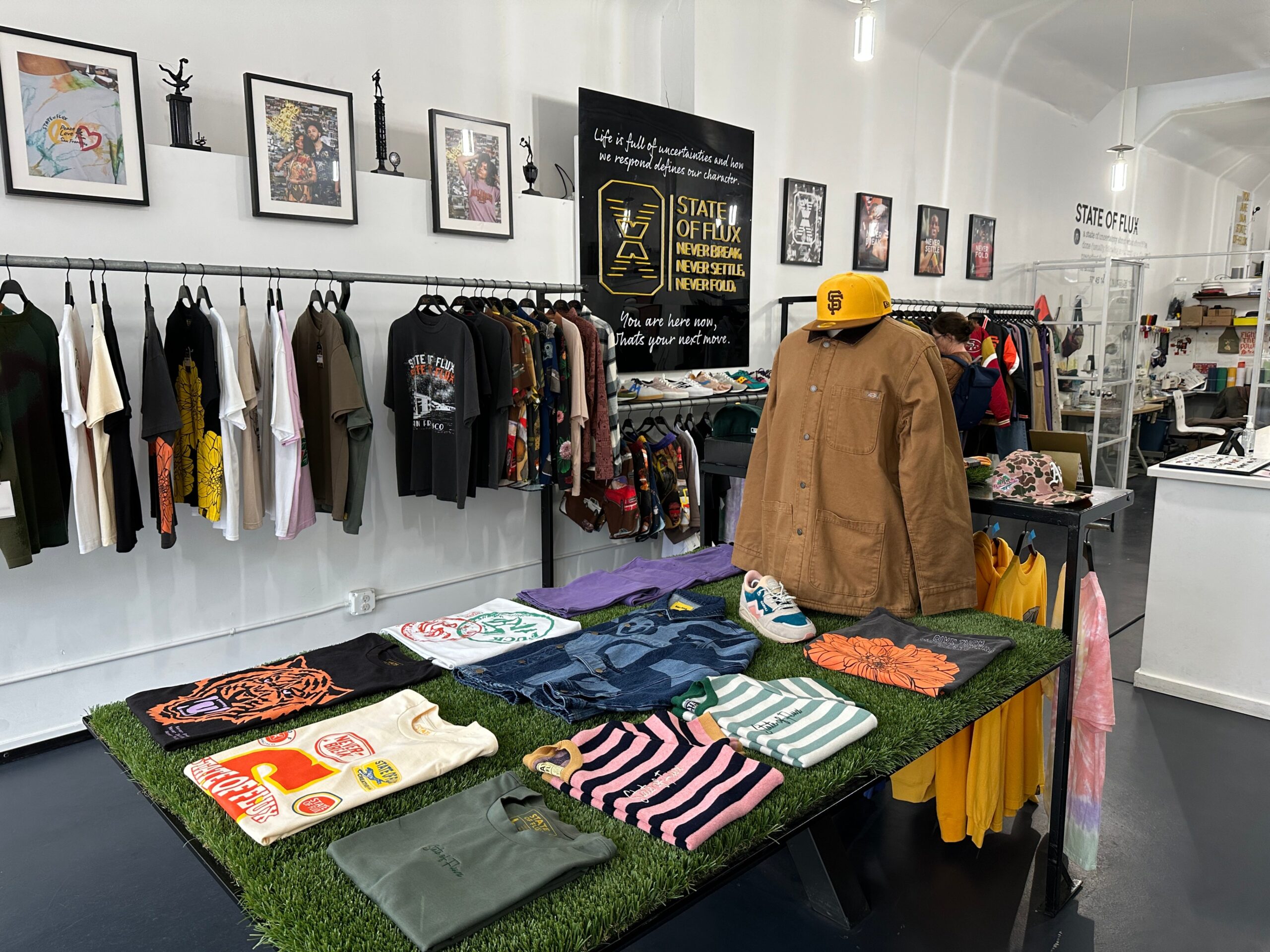 Graphic tees lay on a display in a men's clothing shop filled with racks and a mannequin topped by a yellow SF baseball cap.