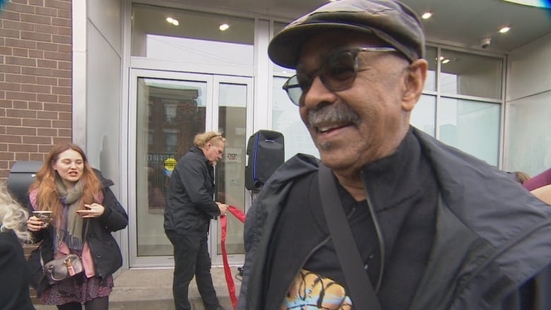 A middle aged man with a mustache talks to a microphone on a city street on a cold grey day. There's a crowd around him, but minding their own business.