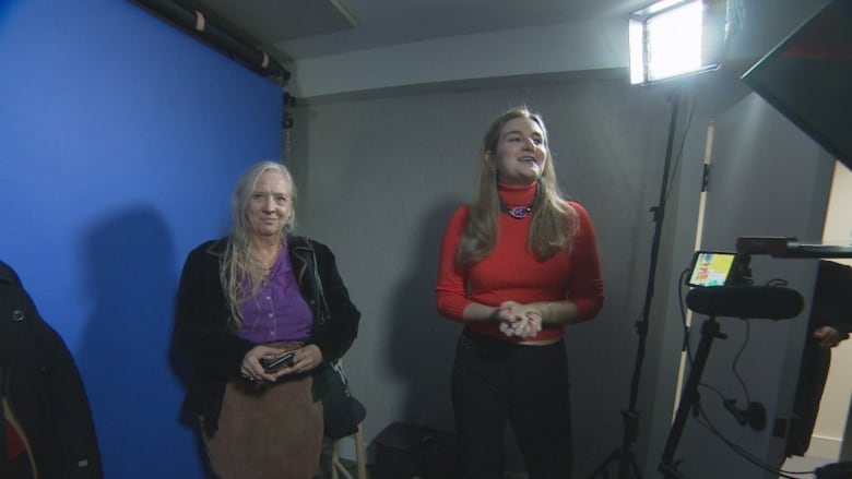 A young woman and middle aged woman talk before a camera and blue screen in a studio.