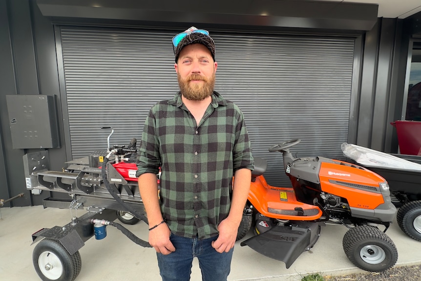 A man in a flannelette shirt and cap stands in front of rural machinery and a scuffed wall