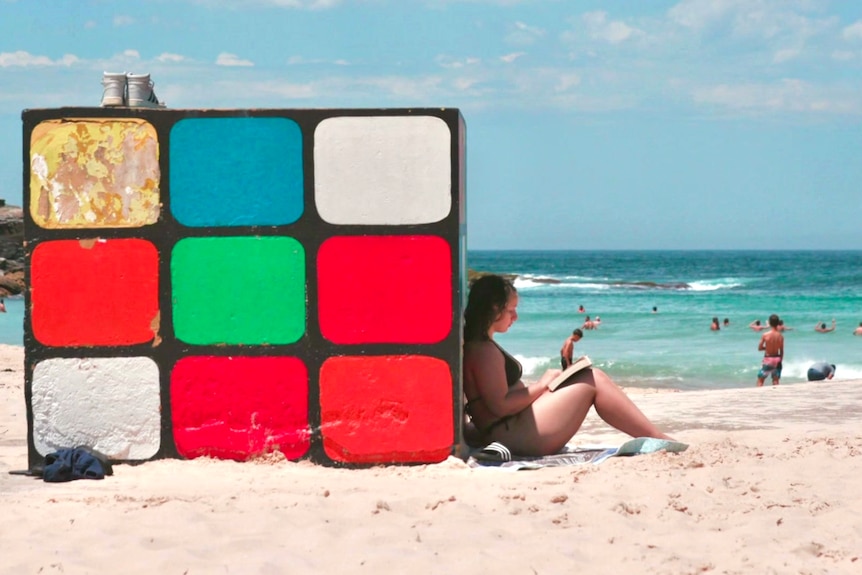 A woman reads a book on the beach while leaning against a giant Rubiks Cube 