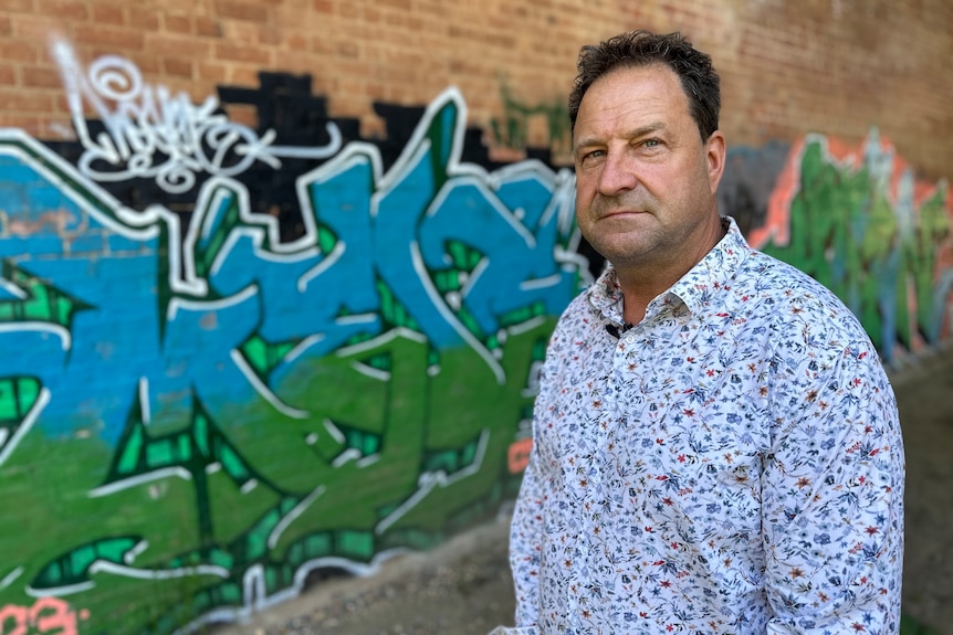 A man in a floral t-shirt stands in front of a graffiti wall