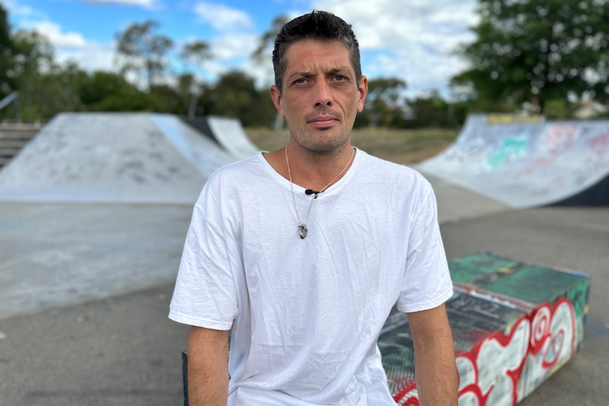 A man with black hair and a white t-shirt looks at the camera
