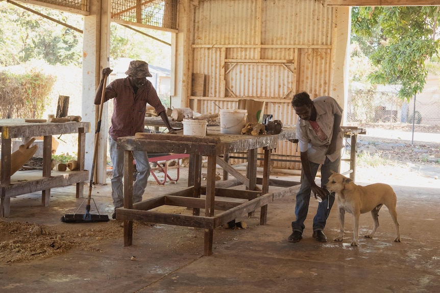 two men stand in a workshop, one feeds a dog from a cup.