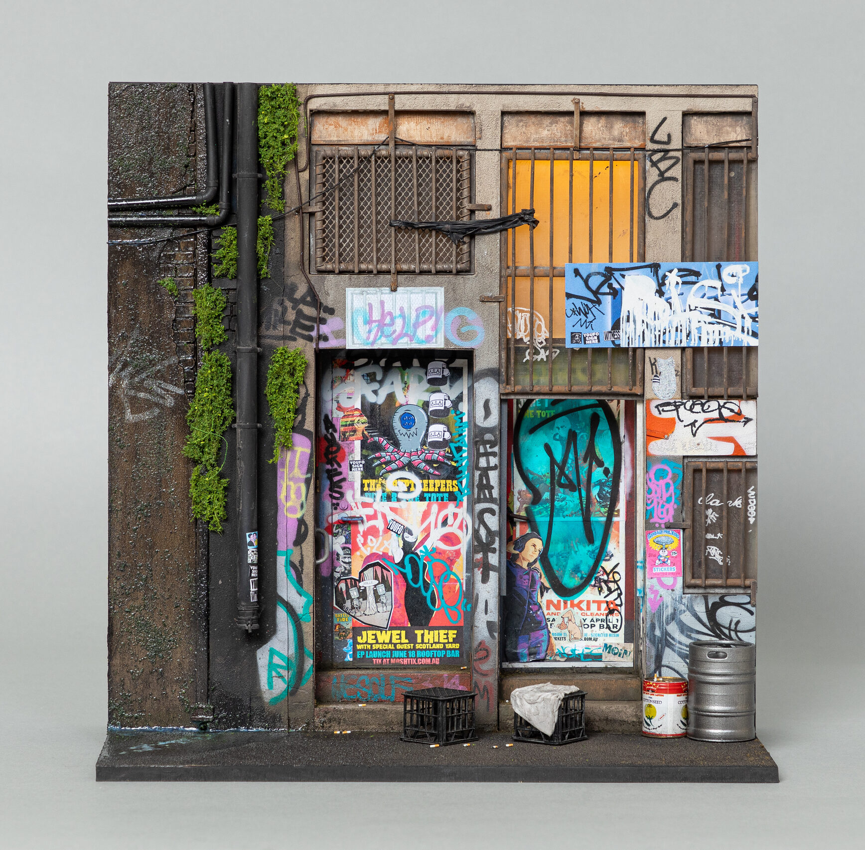 A 1:20 scale model of a building facade with lots of posters and graffiti on the doors, and moss growing near the pipes.