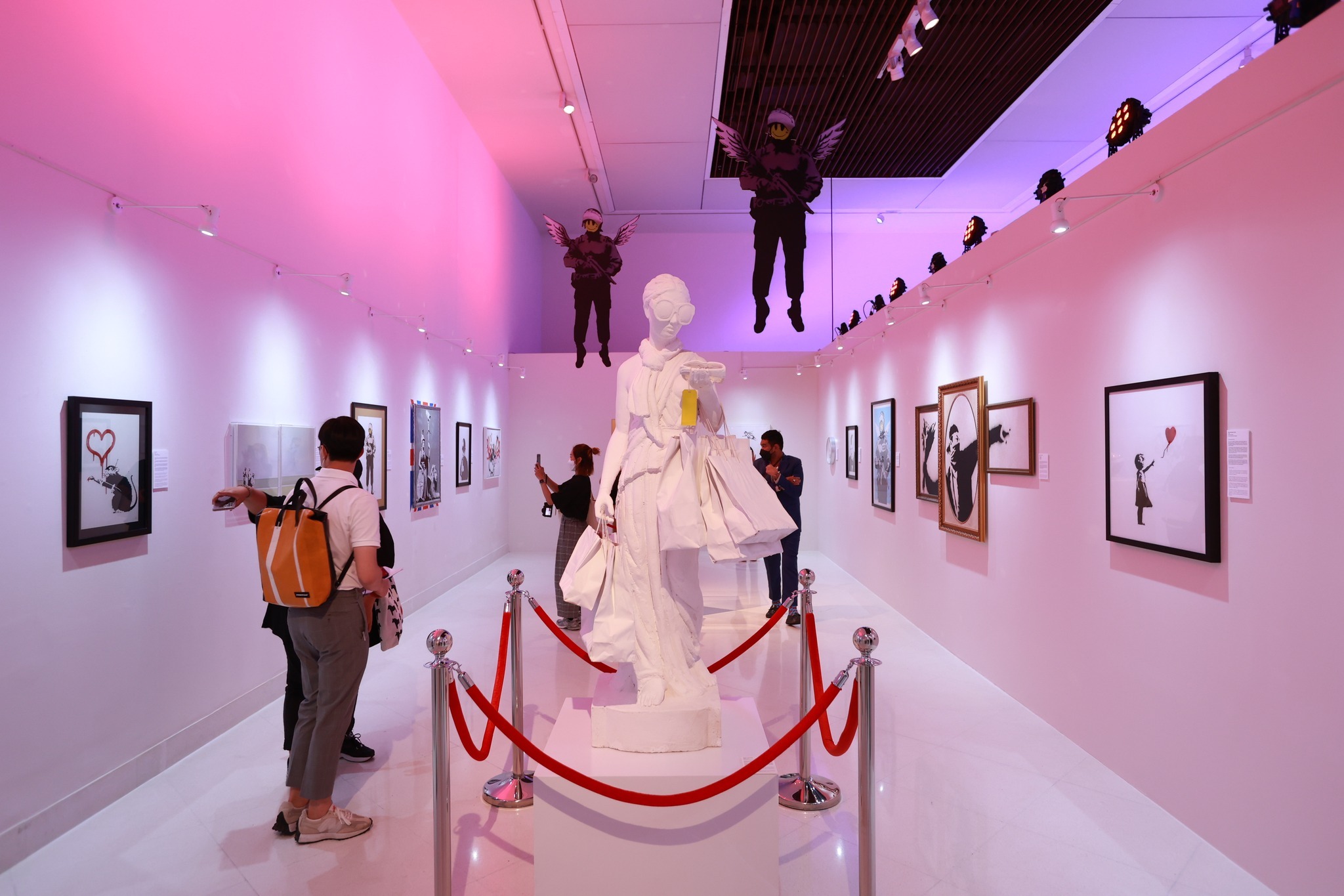 A white sculpture in a purple-lit gallery space filled with Banksy artworks.
