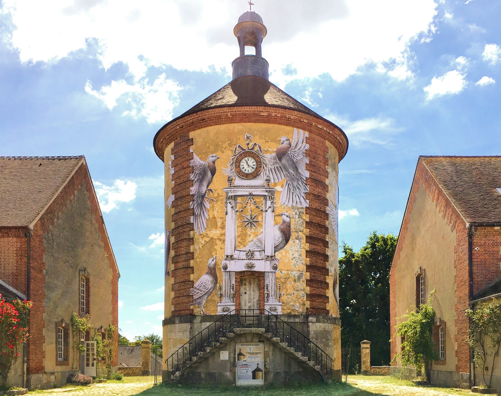 four bird wheatpastes cover a conical structure with a cupola on top in a lush green landscape with brick buildings on either side