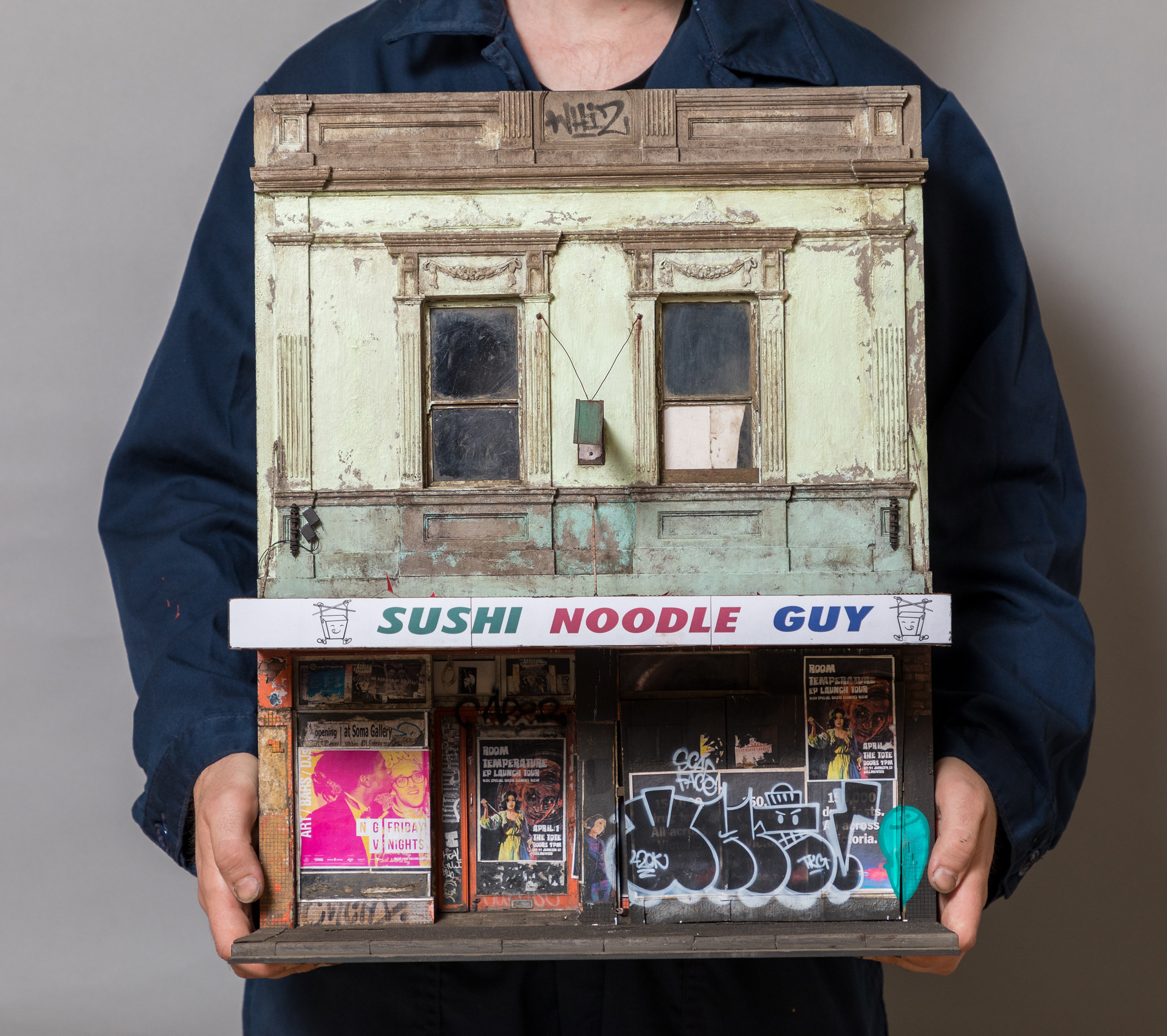 A figure holds a 1:20 scale miniature sculpture of a facade of a shop with posters and graffiti on it.