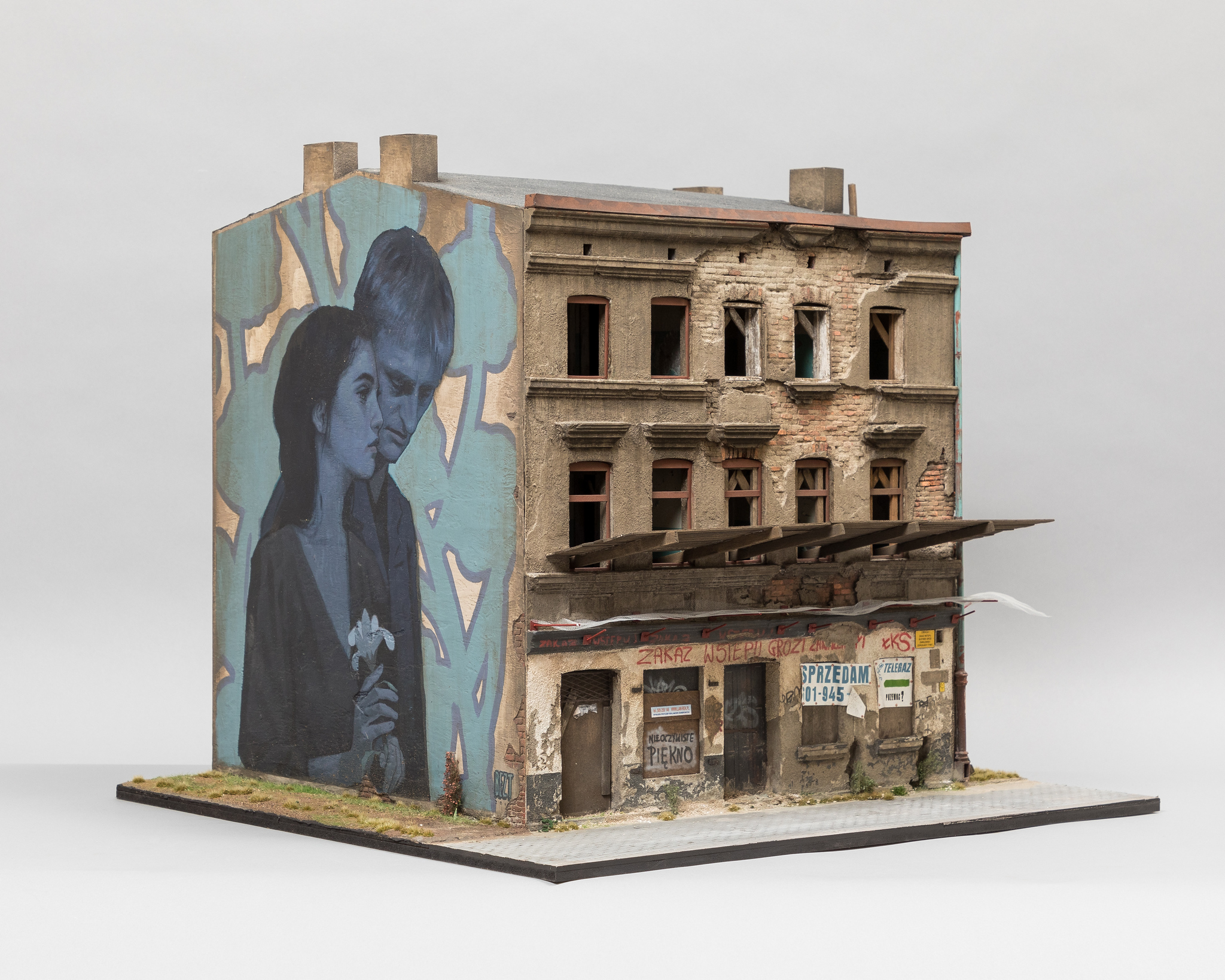 A scale model of an aging apartment building with posters and crumbling signs on the front. On the side, a mural of two figures covers the blank end.