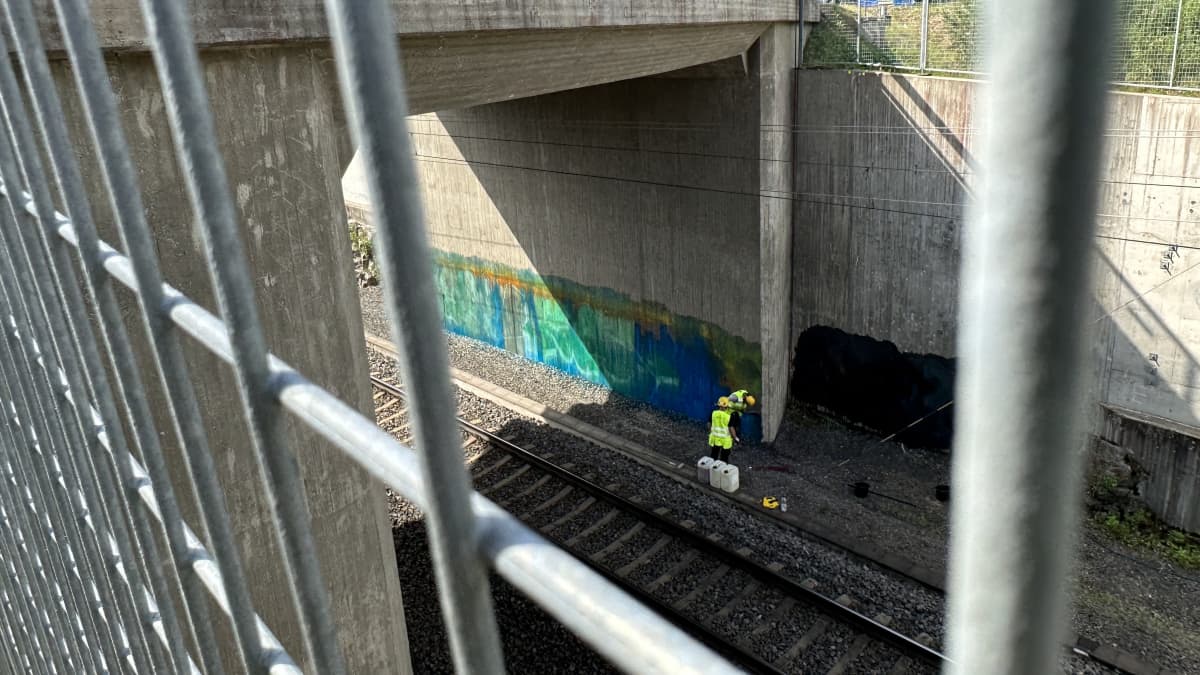 Two city workers wearing bright vests painting over graffiti in a railway underpass.