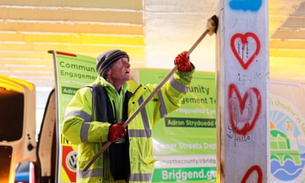 An older man wearing a hi-vis jacket and a beanie hat scrubs a pillar with a broom above head height. He is in front of a van labelled ‘Community engagement’.
