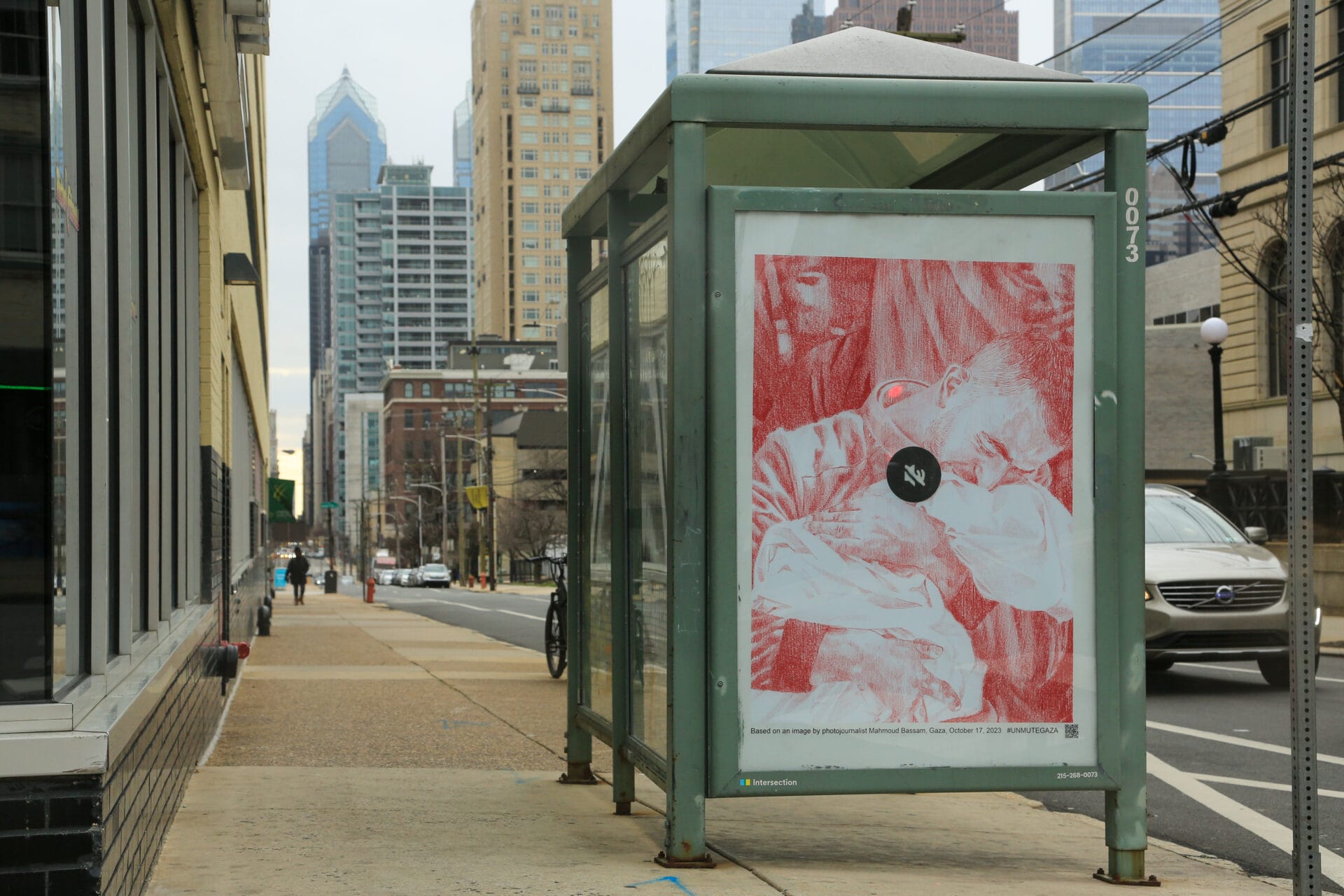 A poster is featured at a bus stop in a city. In the poster, a man clutches onto a baby he has lost, wrapped in white cloth. A mute symbol sits in the middle of the composition. 