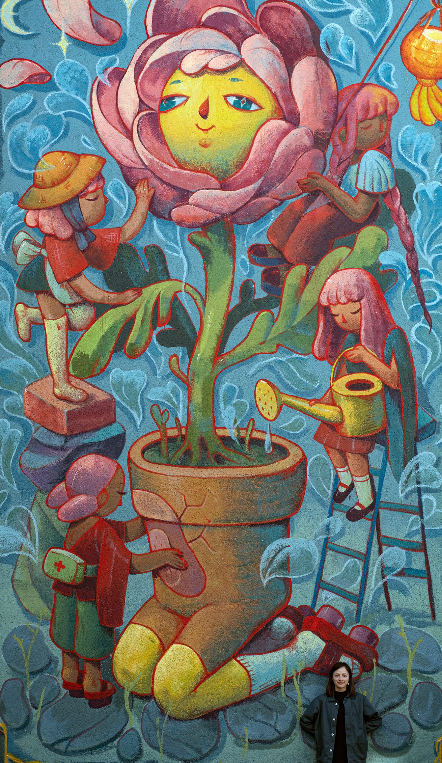 The artist, Marija Tiurina, stands at the base of a tall mural depicting a flower with a smiling face in a pot, with gardeners attending to it.