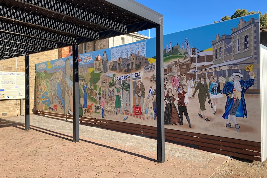 A colourful mural of a town on a wall with people in Victorian clothing, a town crier with a bell in his hand, blue robes.