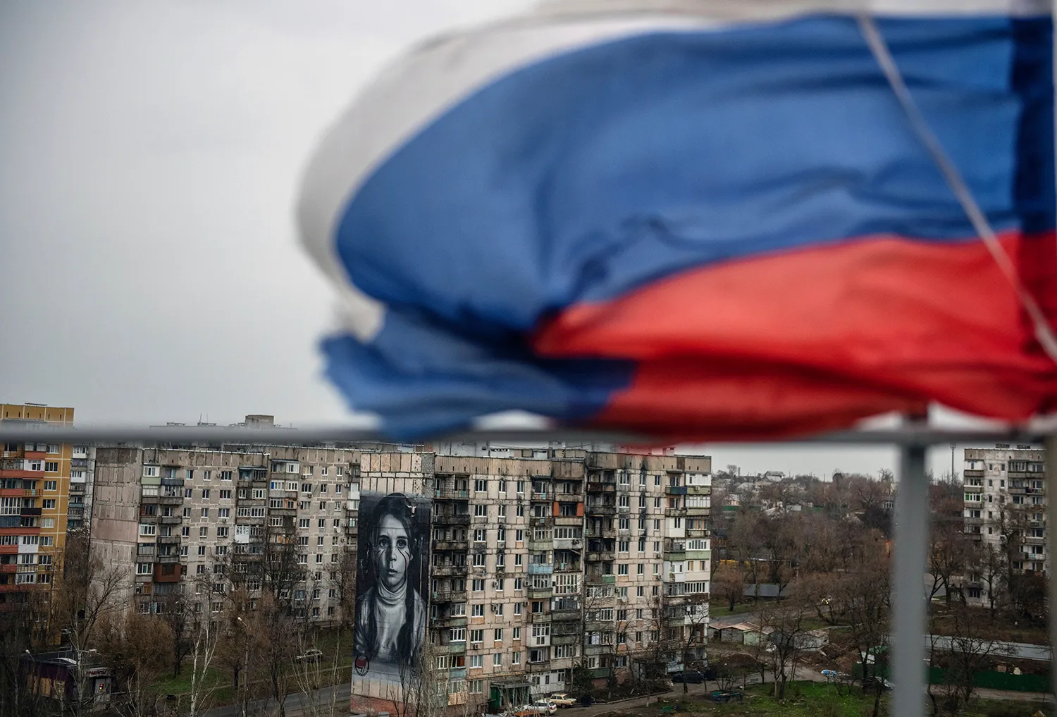 At the top of the image, an out-of-focus close-up of a Russian flag waves in the wind against a white sky. Below, in the background, a worn-down residential building is painted with a large vertical mural of a young girl frowning, with red streaks below her eyes. A few bare trees are visible surrounding the building.