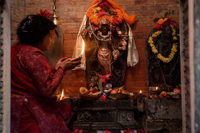 A devotee is seen in profile as she performs a prayer ritual in front of statues of Hindu gods, which are surrounded by candles. The woman, dressed in red, holds an object that has been set on fire while she kneels. Her eyes are closed as she prays.