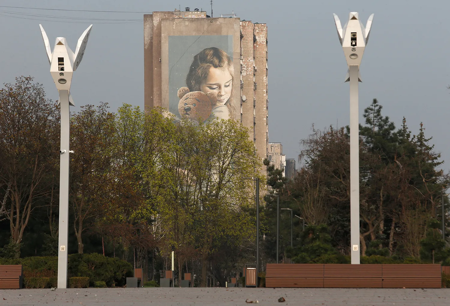 From ground level, two parallel street lights stand on either side of a corridor of trees leading to the background. Where, a tall brick building has a vertical mural of a young girl holding a teddy bear overlooking the scene.