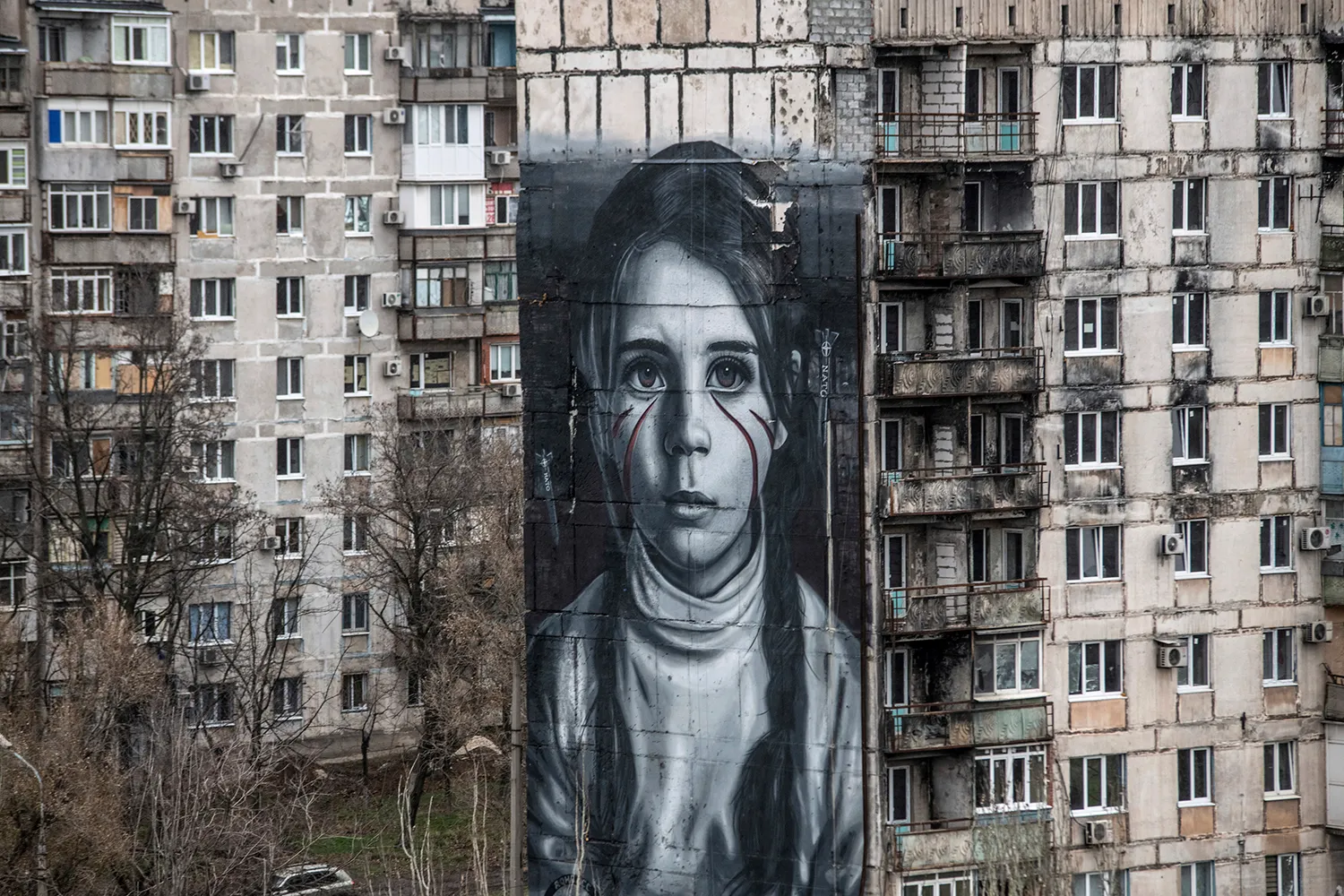 Two weathered residential building stand side-by-side, with many windows and balconies facing the camera. In the middle third of the composition, a large mural of a girl with red streaks below her eyes is depicted on the narrowest side wall of one of the buildings, at a slight diagonal angle to camera.