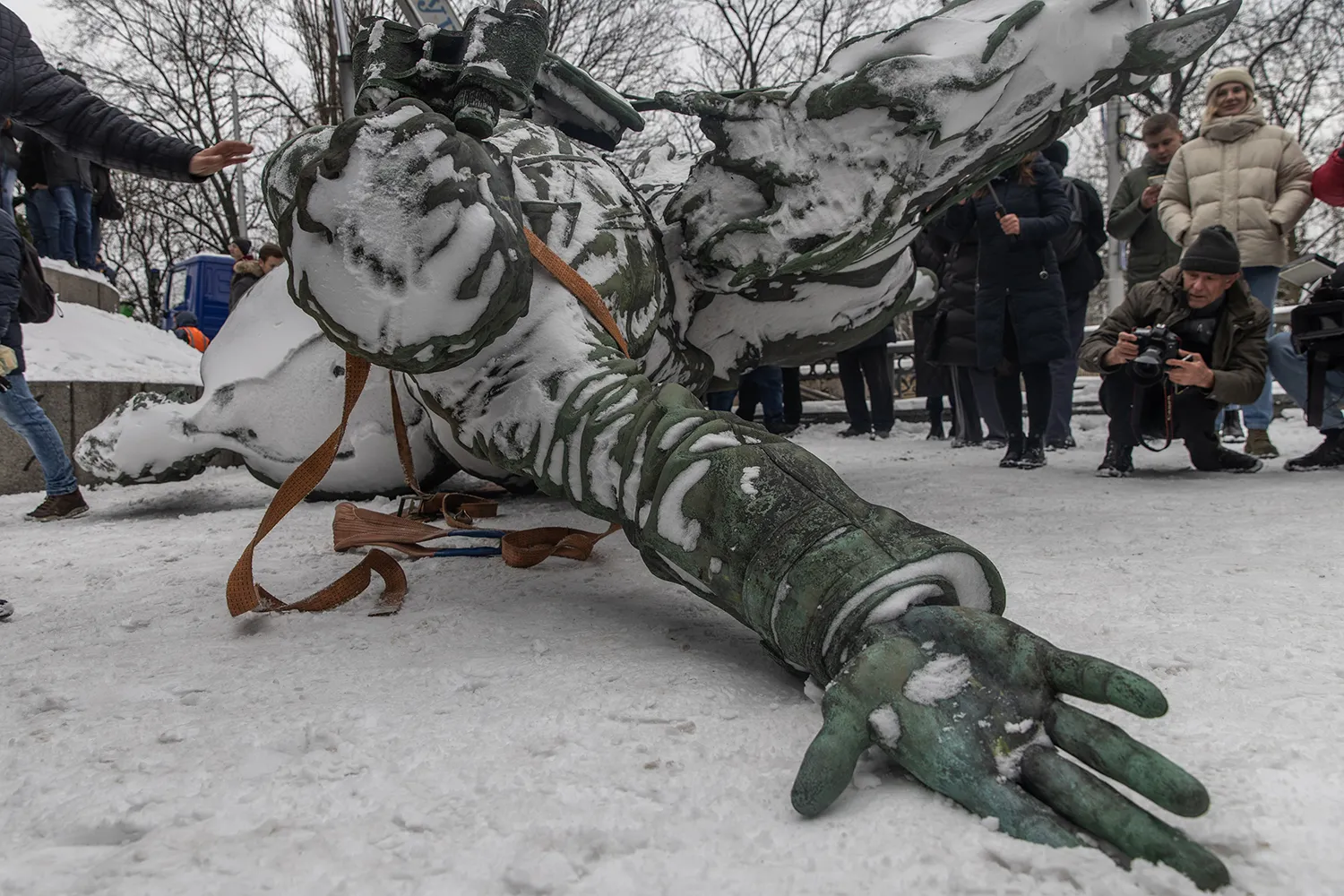 From ground level, a close-up of a fallen bronze statue dominates the frame. There are bystanders and a man crouching to take photos of the snowy scene. The monument lying on its back and covered in snow. The figure's outstretched right arm seems to reach toward camera with an open palm.